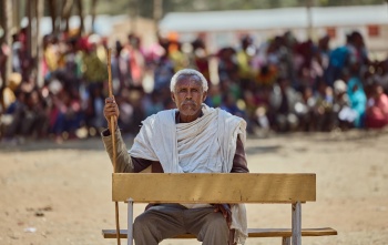 Image of Beyne Bsrat sitting at a desk against the backdrop of a crowd which have gathered.