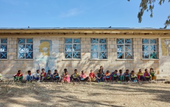 Children sitting outside of their school in a line enjoying their school meal. 