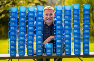 Magnus MacFarlane-Barrow smiling at the camera surrounded by Mary's Meals blue mugs. 