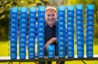Magnus MacFarlane-Barrow smiling at the camera surrounded by Mary's Meals blue mugs.