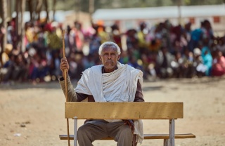 Image of Beyne Bsrat sitting at a desk against the backdrop of a crowd which have gathered.