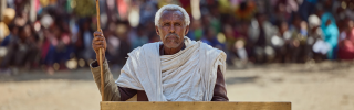 Picture of Beyne Bsrat sitting a desk looking into camera, against the backdrop of an out of focus crowd which has gathered.