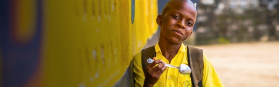 A boy leans against a yellow wall as he eats a bowl of food