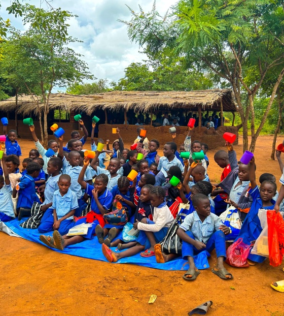 Children at school sitting outside with their cups for their daily school meal.