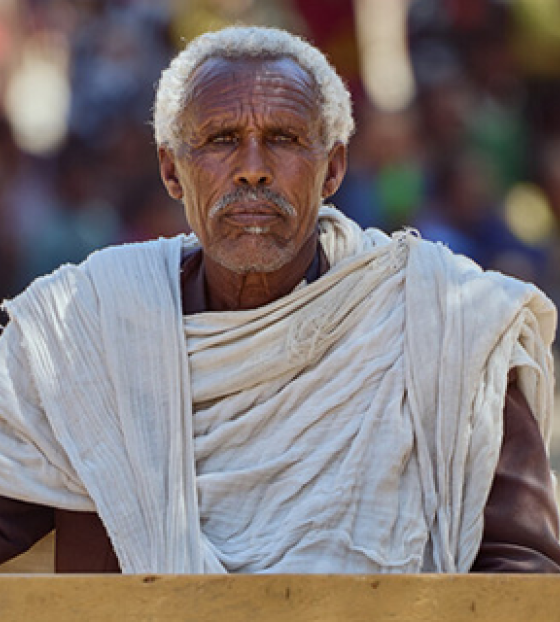 Picture of Beyne Bsrat sitting a desk looking into camera, against the backdrop of an out of focus crowd which has gathered.