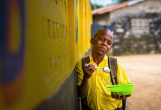 A boy leans against a yellow wall as he eats a bowl of food