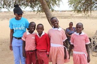 Children standing together with a member of the Mary's Meals team smiling and looking into camera outside against a desert backdrop. 