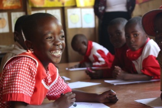 Image of a young girl in school uniform smiling, looking away from the camera. Other children can be seen in the background working on school work. 