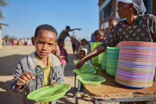A picture of a boy holding a plate and spoon ready to receive a school meal.